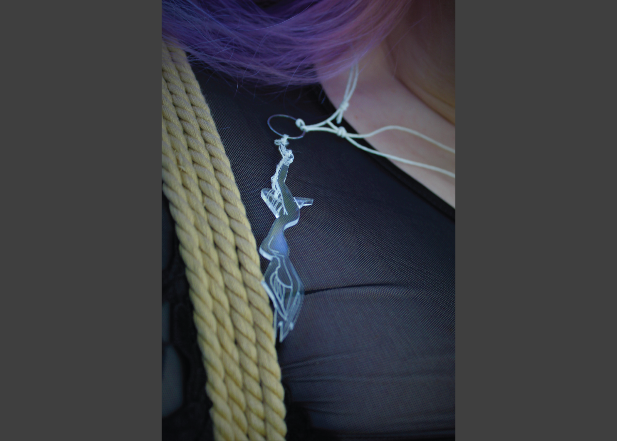 Close up of an acrylic necklace, laser engraved, cut like a women in a self-suspension. Woman necklace piece tied in a futo and gravity boot hanging from her legs, arms and hair falling limp above her.