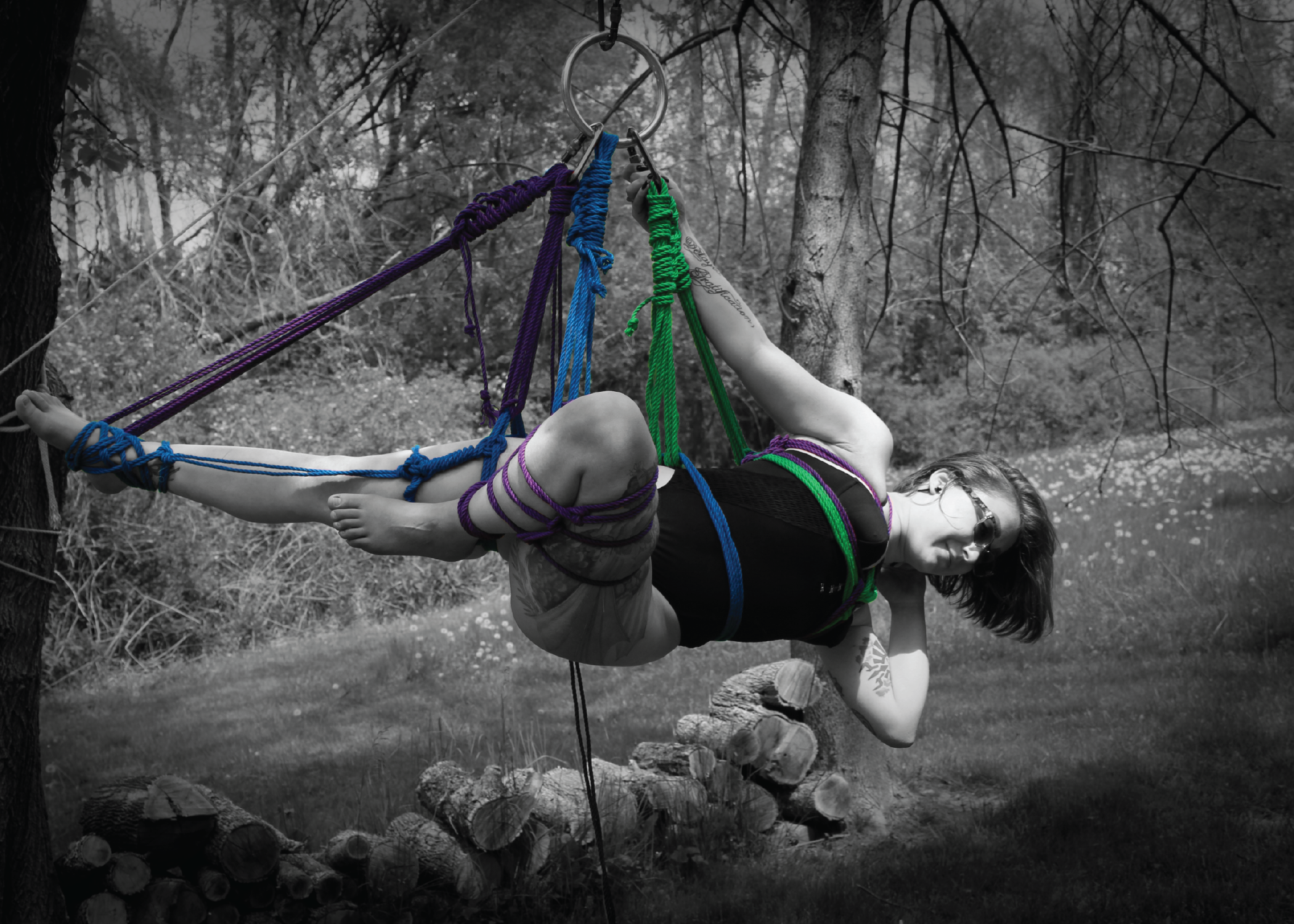 Black and White Color pop Photo of a Beautiful Woman with short dark hair, and sunglasses, Suspended from a tree outside, hands free, posing for the camera. Suspension lines purple & Green, tied in a chest harness, gravity boot on extended leg attached to hip harness on top leg, futo on lower leg.