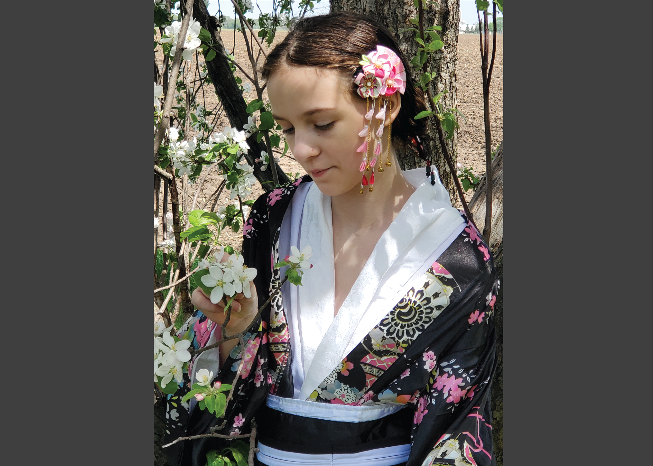 Portrait of a fairskinned girl in japanese garb under a flowering tree looking at a white blossom.