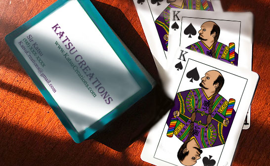 Image of a split deck of cards on a table. One side with the business information for Katsu Creations, the otherside with a custom drawn King of Spades holding a rainbow paddle.